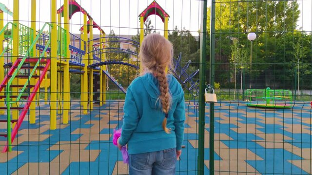 Sad lonely child near the closed playground because of the quarantine.A girl with long blonde braided hair staying alone near empty fenced showground without children.COVID-19 lockdown social distance