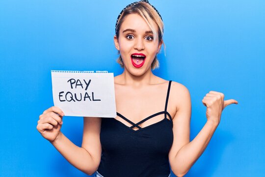 Young beautiful blonde woman holding pay equal paper pointing thumb up to the side smiling happy with open mouth