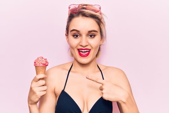 Young beautiful blonde woman wearing bikini holding ice cream smiling happy pointing with hand and finger