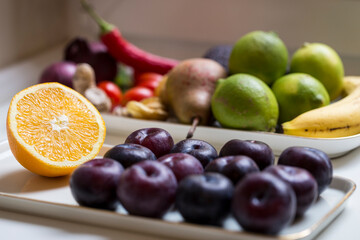 Still life of fruits and vegetables, featuring a sliced orange and several plumbs laid out on plates, lit by lateral soft widow light from a cloudy day