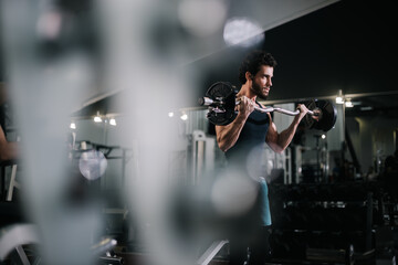 Athletic bearded young man with muscular wiry body wearing sportswear lifting barbell during sport workout training in modern dark gym. Concept of healthy lifestyle.