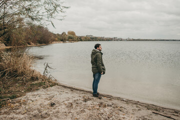 Young sad man in a green jacket walks in nature near the lake in autumn. Traveling, cold weather, rain, thought, thoughtfulness and loneliness.