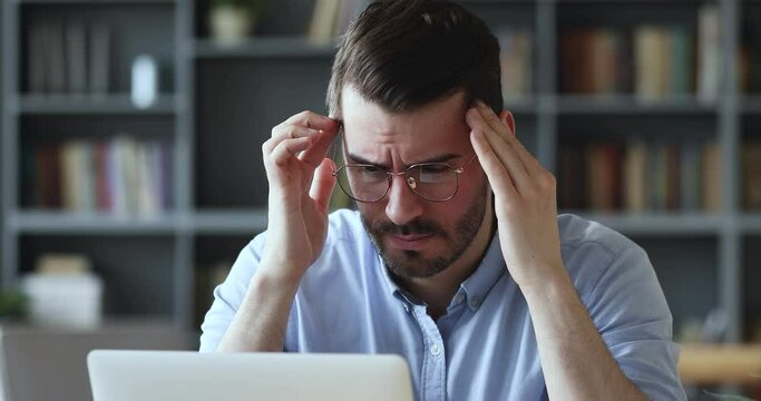 Stressed young man in glasses suffering from muscles tension, having painful head feelings due to computer overwork or sedentary working lifestyle. Tired employee overwhelmed with tasks in office.