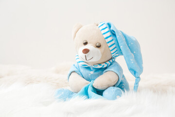 Sleepy teddy bear in blue pajama and hat sitting on white fluffy fur blanket. Kids best friend for sweet dreams. Front view. Closeup.