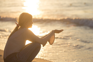 Calm and peaceful young woman sitting alone on the beach sand during a beautiful sunset reflecting on water. Relaxing concept.
