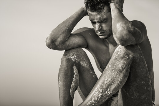 Stressed ill male athlete sitting at the beach with hands on his head. Black and white man portrait.
