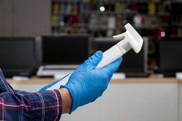 A man in blue protective gloves holds a bottle with a disinfectant in the background of the interior of a computer store. Protection from bacteria and viruses.