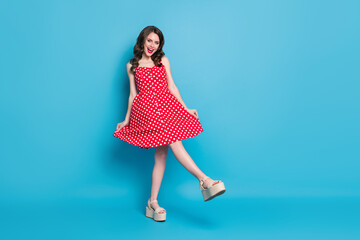 Full body photo of attractive lady shiny pomade smiling toothy flirty hold skirt playful mood raise leg wear red white dotted retro dress summer open toe shoes isolated blue color background