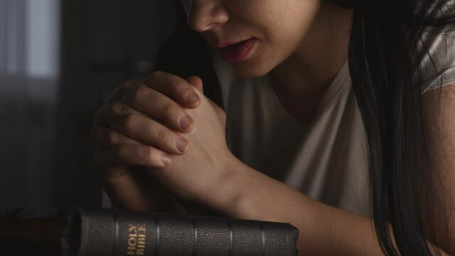 A woman prays with her hands crossed over a Bible for God's blessing in a dark room.