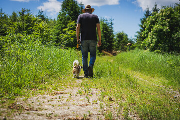 Naklejka na ściany i meble Mann über 40 mit einem Sonnenhut spaziert mit einem kleinen Terrier Hund an der Leine über einen Feldweg im Wald, Einsamkeit, Stille