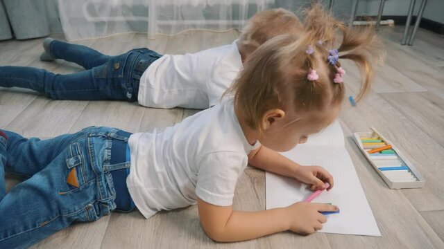 Cute children draw with crayons on white paper lying on the floor in the room.
