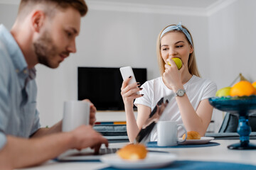 selective focus of girl eating apple and using smartphone near freelancer boyfriend and breakfast