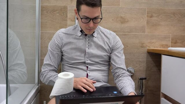 Young handsome Caucasian brunette office worker in plaid gray shirt wearing black glasses, sits on toilet bowl, use laptop Surfing, internet close up. One man with computer calm and relax in bathroom