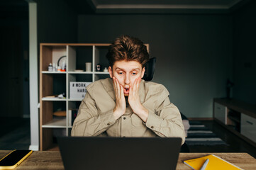 Portrait of shocked young freelancer man working on laptop at home, looking at the screen with...