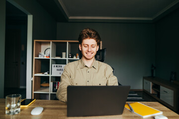 Happy young man in shirt working at home on laptop with smile on face. Joyful freelancer works at home on remote work. The guy uses a laptop on the desktop in the bedroom.