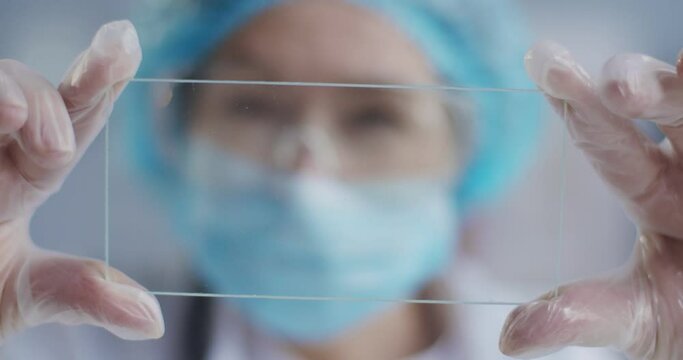 Close up of microscope glass with sample in hands of female Caucasian epidemiologist in mask, goggles and gloves. Lab assistant looking at sample. Laboratory testing. Clinic research. Analysis concept