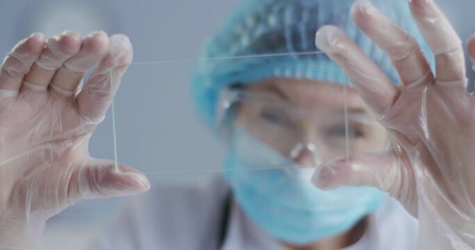 Close up of female Caucasian epidemiologist in mask, goggles and gloves holding microscope glass with sample in hands. Lab assistant looking at sample. Laboratory testing. Clinic research concept
