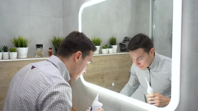 Young Caucasian handsome brunette man wearing gray plaid shirt, looks in mirror uses an irrigator - electric brush for cleaning his mouth ant teeth. Guy wash the oral cavity and spits. Morning routine