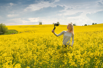 young beautiful blonde girl films herself using a Steadicam in a rapeseed field on a Sunny bright day