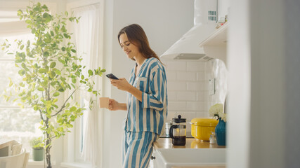 Beautiful Female is Using a Smartphone in a Kitchen While Drinking a Cup of Freshly Brewed Coffee. Girl in Pyjamas with Healthy Lifestyle Relaxes at Home in the Morning.