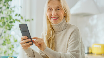 Authentic Senior Woman Using Smartphone in Kitchen Room at Home. Beautiful Old Female Pensioner with Gray Hair Uses an Application on a Cellphone. Elderly Person Looks at Camera and Smiles.