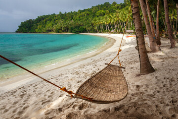 A wicker hammock hangs between two coconut trees against the backdrop of a tropical beach and blue sea.