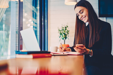 Skilled female copywriter reading news from networks on modern smartphone while doing remote job in...