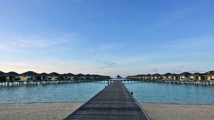 Obraz premium A clear sunny day in the Maldives. A wooden platform goes from the sandy shore to the villas on the water. Calm aquamarine ocean, bright blue sky. Silence and happiness.