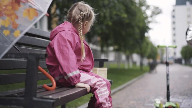 Cute joyful Caucasian girl sitting on bench on rainy day and shaking legs. Side view portrait of little kid in pink costume and rubber boots resting outdoors. Childhood, lifestyle, leisure.