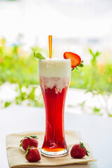 A strawberry ice tea in a tall glass on a white table with strawberry fruits next to the window in the afternoon with nature background.