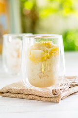 Summer appetizer of Thailand concept. Durian and sticky rice with coconut milk cream in transparent glass on white wooden table and brown table cloth and yellow and green bokeh light background. 