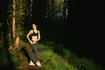 Obraz na płótnie Canvas A beautiful woman in leggings and a top stands in the sunlight in the morning in a summer forest