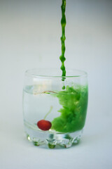 In a transparent glass with clean water, with cherries at the bottom, green paint is poured with a dynamic movement and colors the water green on a white background.