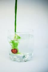 In a transparent glass with clean water, with cherries at the bottom, green paint is poured with a dynamic movement and colors the water green on a white background.
