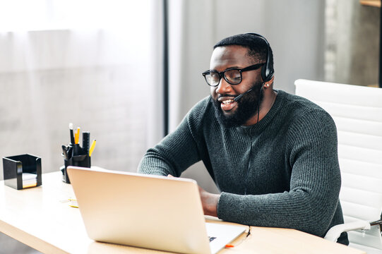 Young African-American Guy Is Call Center Worker Or Support. Smart Black Man In Eyeglasses Uses A Handsfree Headset And Laptop To Talk Online At His Workplace