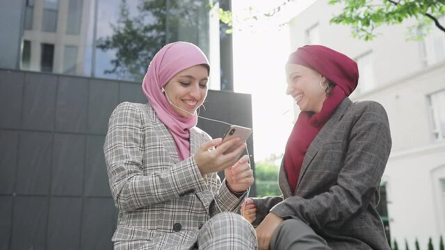 Two young Muslim women wearing hijab headscarf listen music on phone by earphones, sitting together at bench having fun modern lifestyle, technology, smartphone usage, leisure time.