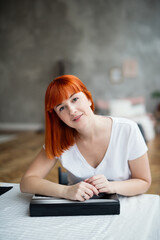 a girl with red hair sits at  table, hands on a book,  white tablecloth, a gray wall