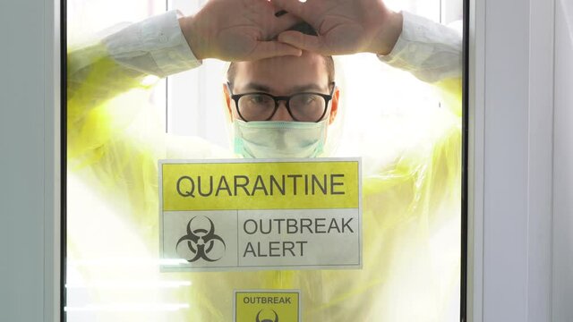 Isolated illness man wear yellow protective suit, turquoise disposable medical face mask, black glasses, white shirt come, looks through glass indoors close up. Door sign: quarantine, outbreak alert.