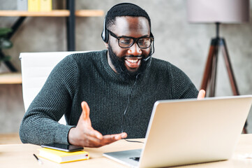 Smiling African-American guy uses a handsfree headset to talk online at his workplace, black confident man in glasses sits at the office desk and looks at laptop screen