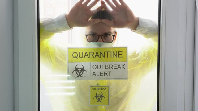 Young serious man with coronavirus infection wear yellow protective suit, medical face mask, stares longingly out window, lean hands, pushes against glass. Isolation by pandemic. Quarantine zone.