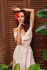 Fototapeta premium Graceful Asian woman with hibiscus flower in hairs posing over wood wall. Tropical leaves. Summer style. 