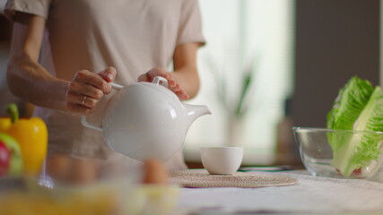 Woman pouring tea into cup from teapot. Lady preparing tea for breakfast at home