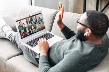 Video call concept, virtual meeting. Young African-American guy relaxed lays on the couch and is...
