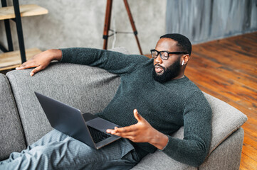 Serious African American guy with glasses lies on the couch and using laptop for online talking via...
