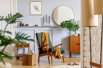 Trendy interior of living room with design armchair, vintage commode, round mirror, mock up poster...