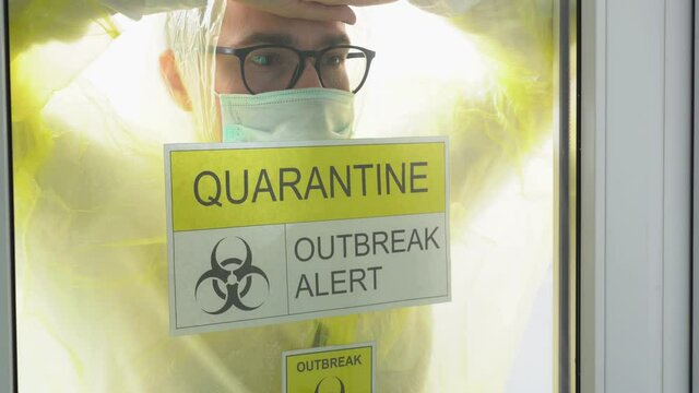 One tired man wear yellow protective medical suit, black glasses, face mask come up to window, leans on glass, looks straight close up. Prohibition sign: quarantine, outbreak alert. Isolation concept