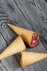 Strawberry ice cream in a waffle cone. Garnished with Chocolate. Nearby are empty waffle cones without ice cream. On pine boards painted in black and white.