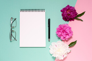 Stylized female office desktop with blank notepad, glasses and bright peony flowers on two tone background top view. Back to office or back to school concept.
