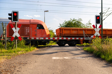 Closed barrier at a railway crossing with red warning lights on, a blurred locomotive in motion is pushing wagons.