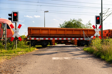 Closed barrier at a railway crossing with red warning lights on, a blurred locomotive in motion is pushing wagons.